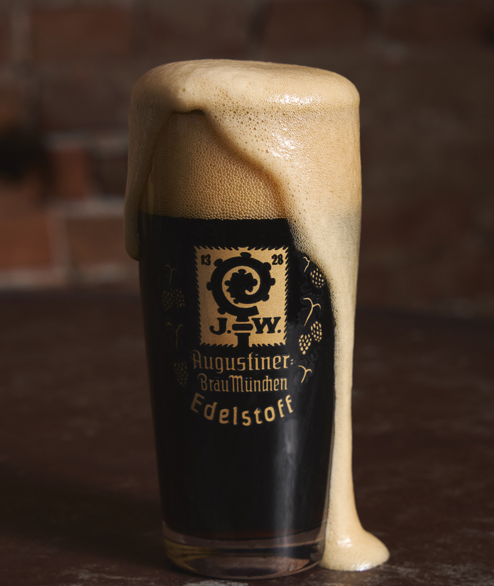 A glass of dark beer with a thick, creamy head, labeled "Edelstoff," with Augustiner Bräu München branding.