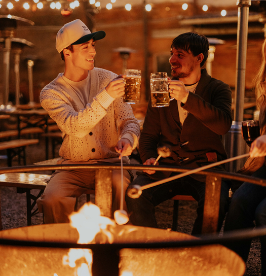 Two men toasting with drinks at a restaurant, surrounded by a warm fire pit and outdoor lights.