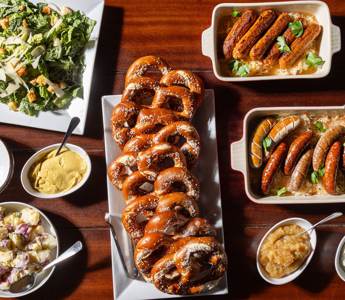 Assorted food items including pretzels, sausages with sauce, Caesar salad, potato salad, and mustard on a wooden table.