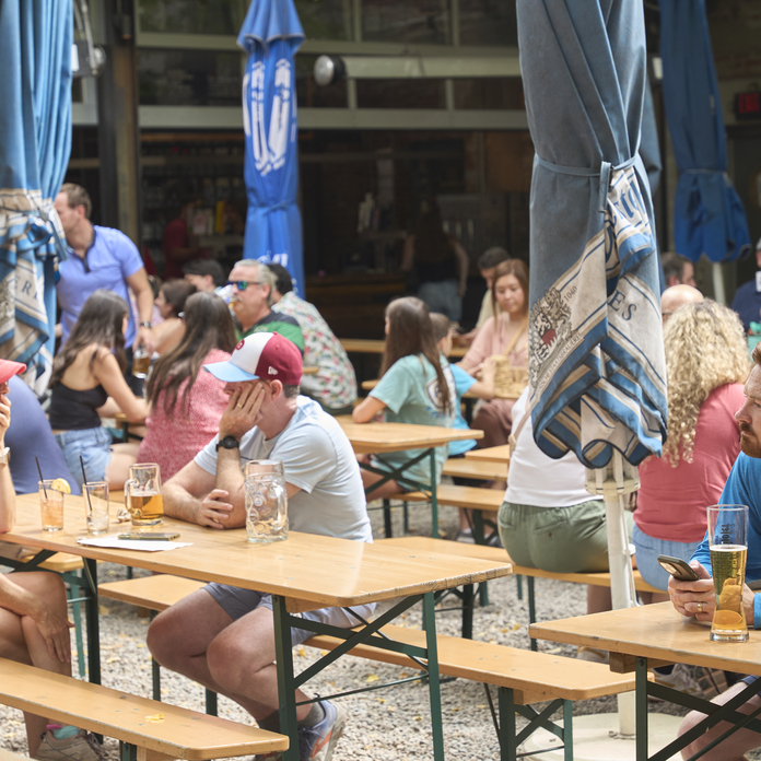 People enjoying outdoor seating at a restaurant with wooden tables, metal legs, and blue umbrellas.