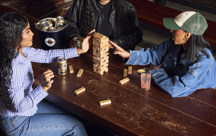 Three women play Jenga at a rustic wooden table in a cozy restaurant setting.