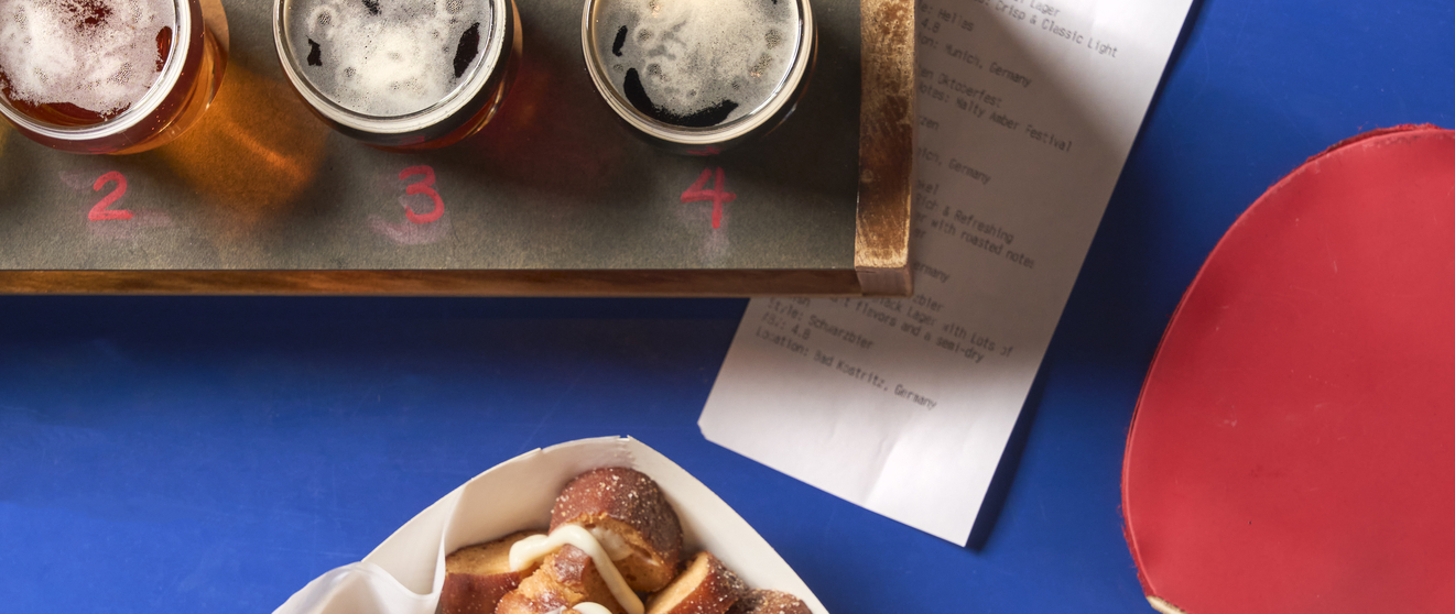 Overhead view of a restaurant table with a loaded sandwich, beer flight with glasses, pretzels, ping pong paddles, and an order slip.