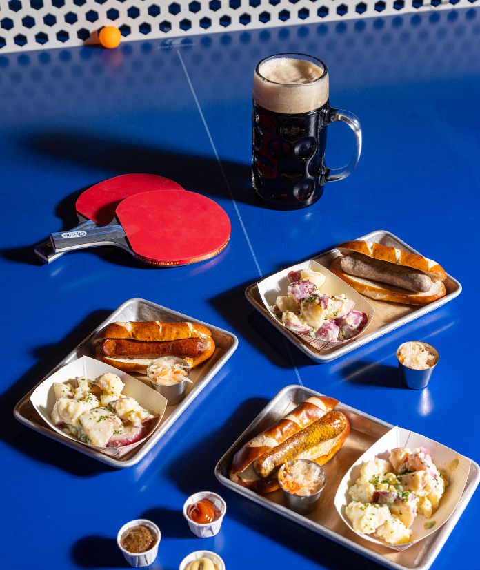 An overhead view of a blue ping-pong table set with metal trays of sausages, lobster rolls, potato salad, and condiments alongside two red paddles, a ping-pong ball, and a large dark beer stein.