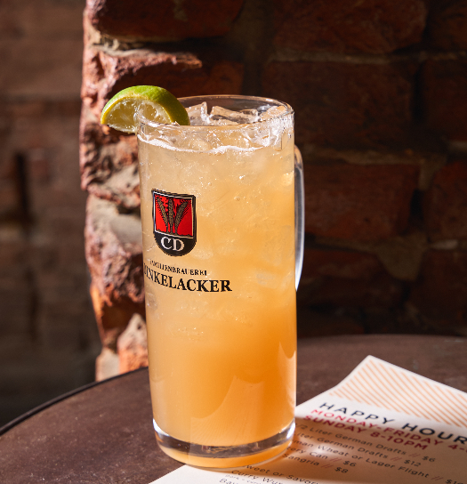A branded Zwickelbacker beer glass filled with a golden, ice-cold cocktail garnished with a lime wedge, set against an exposed brick wall with a happy hour menu visible in the background.