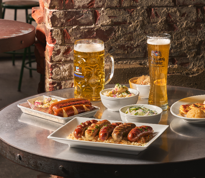 A German-style beer hall spread on a metal table featuring a platter of grilled sausages over sauerkraut, side dishes of potato and cucumber salads, a large Hofbräu beer stein, and a tall pilsner glass, set against an exposed brick wall.