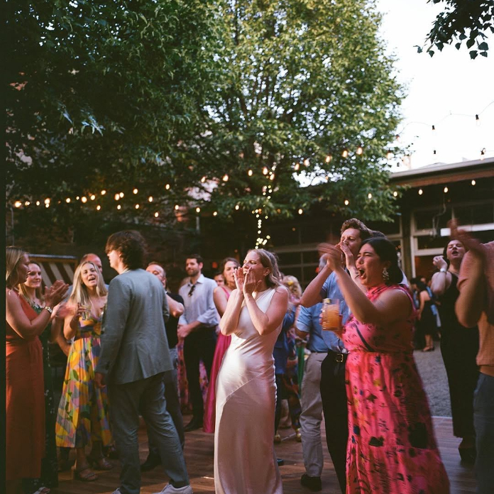 A lively outdoor wedding reception with guests dancing and clapping on a wooden dance floor, surrounded by string lights and lush trees at dusk, with a woman in a white gown at the center.