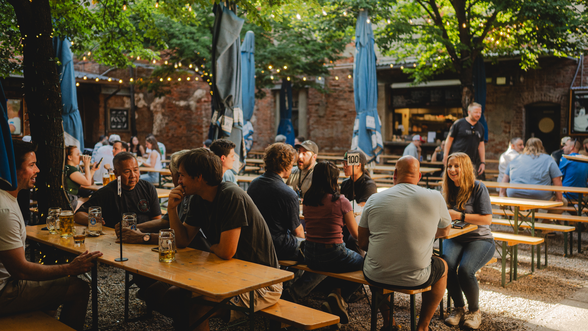 Outdoor beer garden with people sitting at wooden picnic tables under trees with string lights and brick buildings