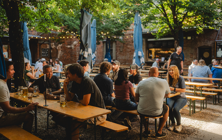 Outdoor beer garden with people sitting at wooden picnic tables under trees with string lights and brick buildings