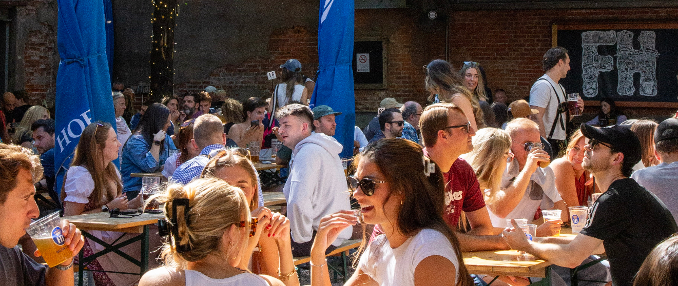 Crowded outdoor beer garden with people seated at picnic tables enjoying drinks under blue umbrellas against brick wall